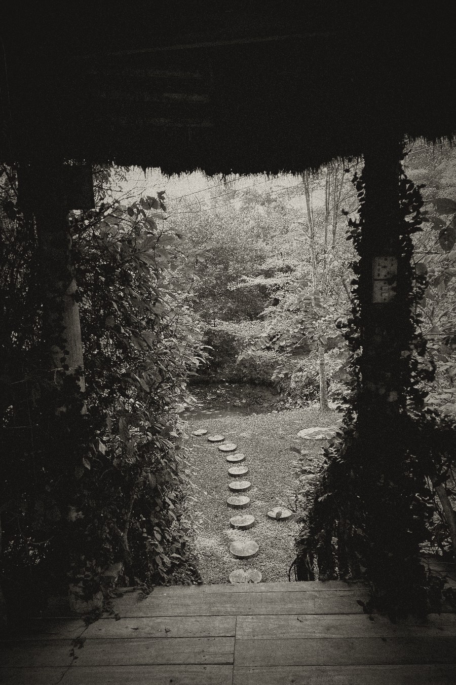 Desaturated charcoal print of a view from a rustic thatched hut onto a shadowed path of stepping stones